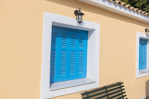 Vibrant blue wooden shutters frame a window on a traditional Mediterranean building with warm beige walls and classic lantern. Authentic Greek island architecture.