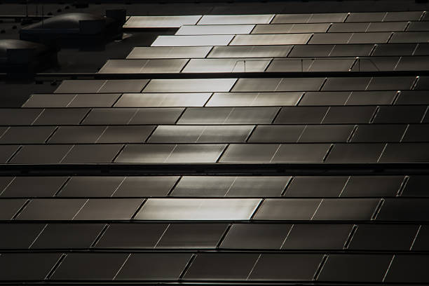Close-up view of solar roof tiles designed to resemble traditional roofing materials. The tiles are arranged in a uniform, overlapping pattern and feature a glossy surface that reflects sunlight. The image highlights sustainable energy integration.