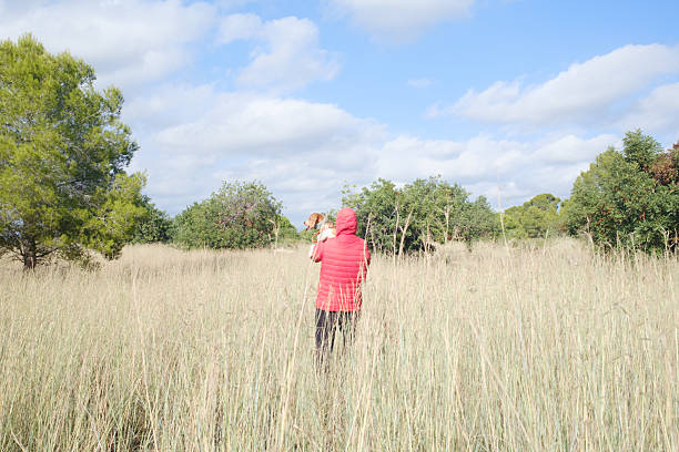 Person wearing red jacket carrying a small dog while walking through a vast field of dry tall grass under a blue sky with scattered clouds, exploring nature
