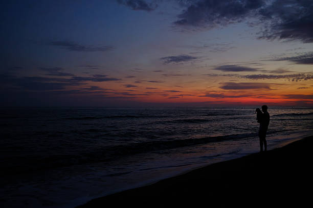 Man holding small dog at water’s edge, silhouetted against a colorful dusk sky as gentle waves roll onto a tranquil, golden-orange seaside horizon
