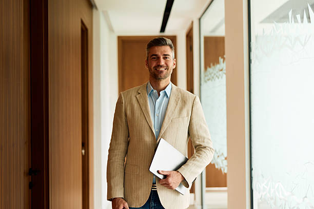 istock Smiling businessman holding laptop in modern office corridor 2239333944