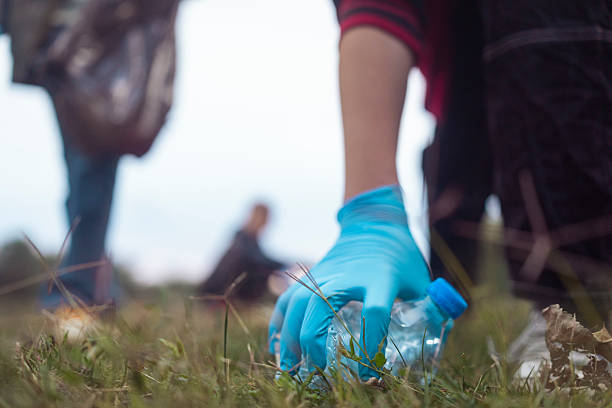 A person wearing a blue glove collects a plastic bottle from grassy ground outdoors with blurred figures in the background during an environmental cleanup effort.