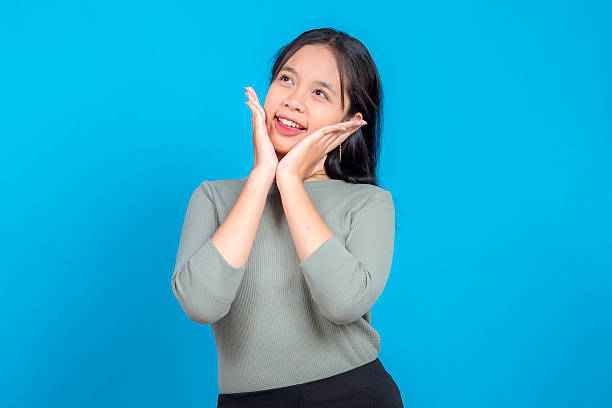 istock Young Asian woman with a joyful expression, smiling brightly and holding her face with both hands in a playful pose while looking up, isolated on a solid blue background. 2239354068