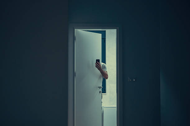 istock Man partially hidden behind door holding smartphone in dimly lit interior, mysterious atmosphere. Hand with smartphone visible through open door, moody low light interior scene with suspenseful feel. 2239356844
