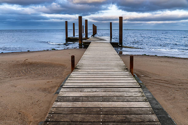 istock Wooden Pier Extends Into Quiet Ocean On Sandy Beach Under Cloudy Sky During Gloomy Evening, Hjerting, Denmark 2239368718