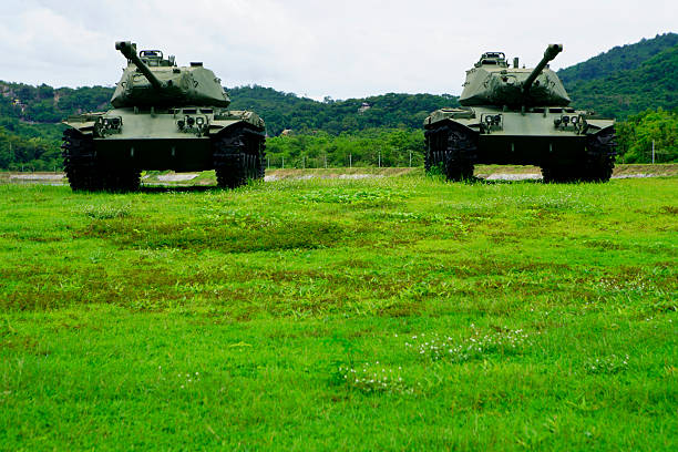 A wide-angle front view of a Japanese self-defense force military tank driving down a dirt road lined with trees with a dramatic sky.