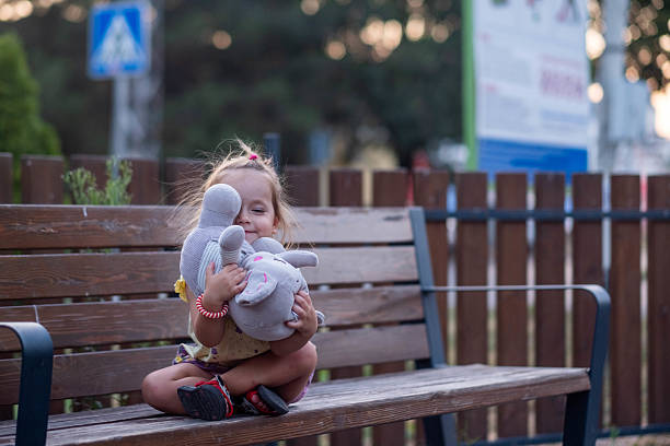 Toddler girl embracing two plush toys, sitting outdoors and smiling happily