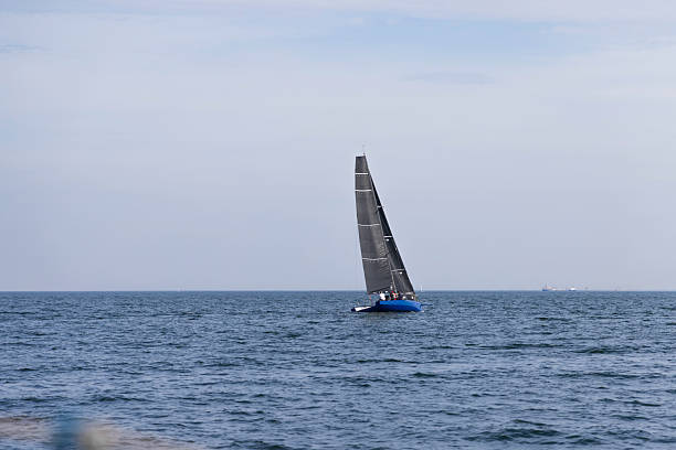 A blue sailboat with tall dark sails glides across a tranquil sea beneath a pale, expansive sky. Ideal for visuals of travel, leisure, adventure, and outdoor recreation.