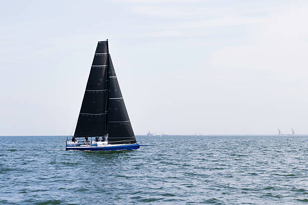 A sleek sailboat with large black sails sails across a blue ocean, crew onboard, distant horizon. Ideal for themes of travel, adventure, freedom, and maritime recreation.