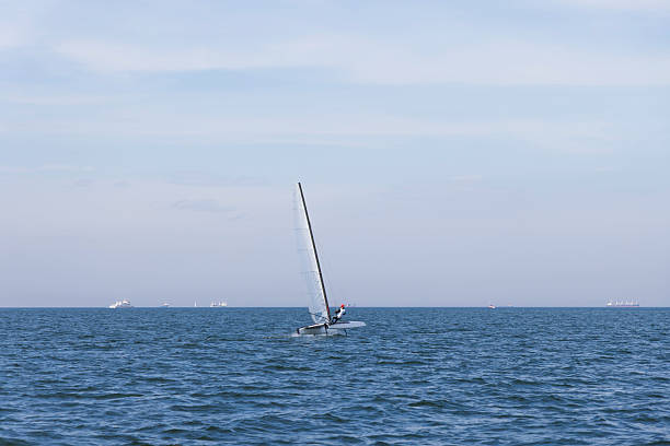 A lone sailboat cuts through calm blue waters beneath a tranquil sky, with several distant ships on the horizon, suggesting travel, leisure, and maritime life.