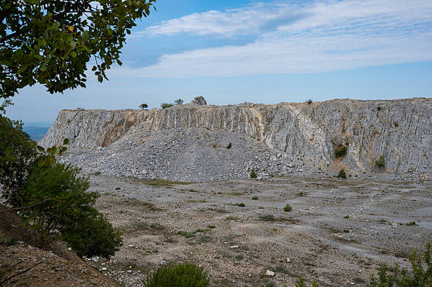 A vertical limestone cliff inside an abandoned quarry, showing weathered rock faces with orange mineral streaks and sparse vegetation. The flat quarry floor is covered with small stones and patches of grass under a bright blue sky with scattered clouds.