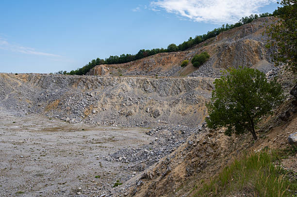 A view of a limestone quarry with steep, layered walls and loose stones scattered across the ground. Sparse green vegetation grows along the slopes and the quarry floor, contrasting with the gray and orange tones of the rock. A bright blue sky with light clouds completes the scene.