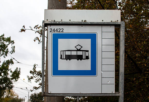 A sign stands at a tram stop, displaying a tram silhouette against a blue background. The surrounding area shows trees and power lines, suggesting an urban atmosphere.