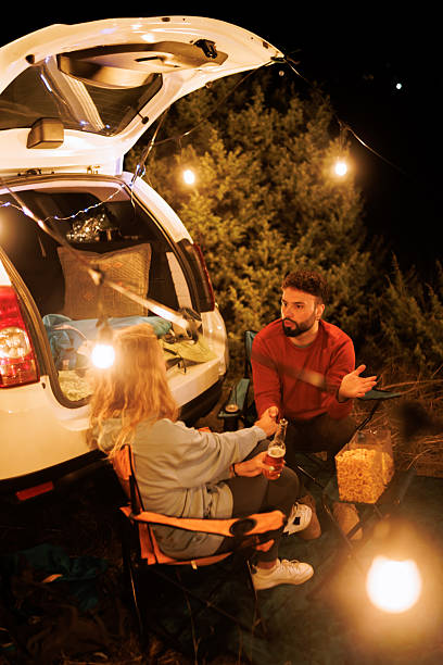 istock Couple shares drinks and stories by their car under string lights at night in a forest setting 2239405087