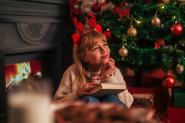 Young girl in reindeer antlers sits by a decorated christmas tree and warm fireplace, reading and daydreaming with a cozy, festive smile of childhood wonder and anticipation