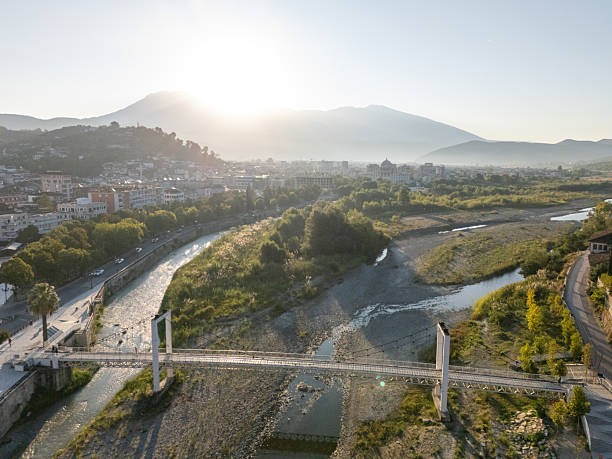A suspension bridge crosses a shallow river with rocky banks in a small town surrounded by green hills and distant mountains. Scenic landscape with urban and natural elements. River flowing through a valley near residential areas.