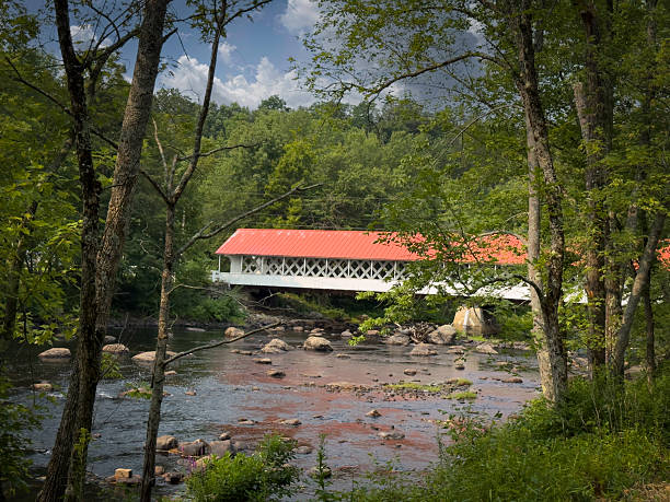 Historic Ashuelot Covered Bridge with a red roof spans the Ashuelot River amid lush summer woods in Winchester, Cheshire County, New Hampshire.
