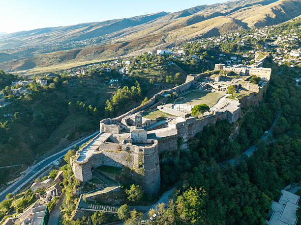 Aerial view of an ancient stone fortress perched on a hill surrounded by lush greenery. Mountain town with clustered houses and winding roads in the background. Scenic historic architecture and nature.