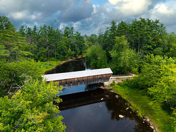 Aerial view of the historic Hemlock Covered Bridge spanning the Old Saco River in Fryeburg, Maine, surrounded by lush summer forest and dramatic clouds.