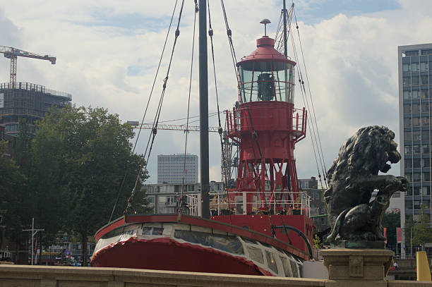 Historic red lightship moored in a modern city harbor with a bronze lion statue in the foreground, blending maritime heritage and contemporary architecture under a cloudy sky.