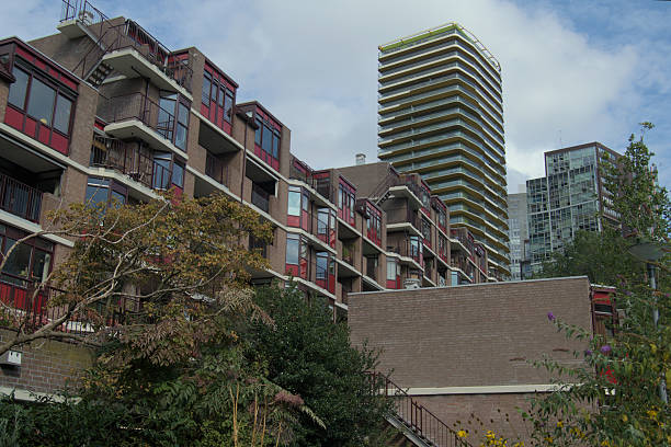 Contemporary residential buildings in Rotterdam, Netherlands, featuring geometric balconies and red window frames. A modern high-rise tower with yellow accents rises in the background, showcasing the city’s blend of architectural styles under a partly cloudy sky.