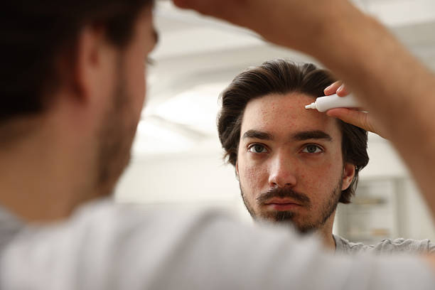 istock Young man with acne problem applying cream onto his face near mirror at home 2239443378