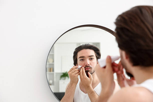 istock Young man with acne problem applying cream onto his face near mirror at home, space for text 2239444237