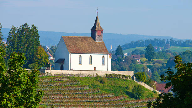 A quaint church stands atop a hill, surrounded by vibrant vineyards and trees. The background features rolling mountains under a clear blue sky, showcasing a peaceful rural scene in Switzerland.