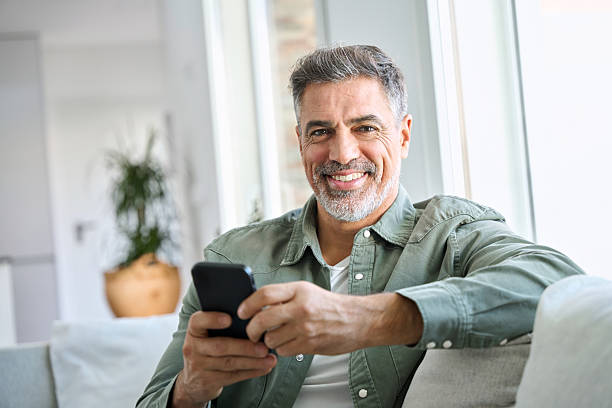 istock Happy mature man using mobile phone sitting at home couch looking at camera. 2239455365