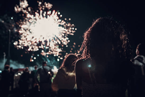 Rear view of a young woman with curly hair looking at bright white fireworks exploding in the night sky. Festive atmosphere with a crowd of people in the background. Concept of celebration and New Year's Eve.