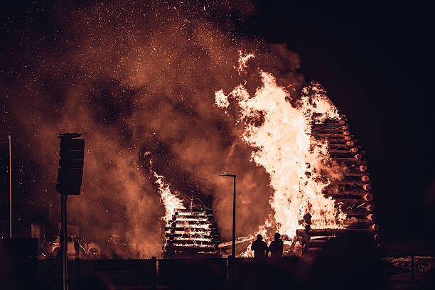 Massive wooden structure burning during a traditional Saint John's Eve celebration (Midsummer festival). Bright orange flames and sparks rising into the dark night sky with silhouettes of people watching in the foreground.
