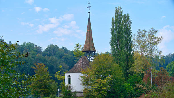 A small, picturesque chapel stands amid vibrant green trees under a bright blue sky. The elegant spire and rustic architecture blend beautifully with nature.