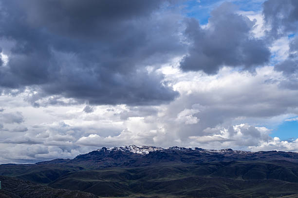 A rugged mountain range in Peru, partially covered with snow, sits beneath a dramatic sky filled with dark storm clouds and patches of blue