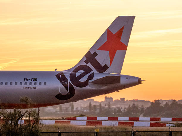 A Jetstar Airbus A320-232 plane, registration VH-VQE, taxiing to the third runway of Sydney Kingsford Smith Airport for departure as flight JQ418 to the Gold Coast. An arrow symbol is built into the logo Jetstar. In the background are residential districts. These sunset colours are caused when strong winds bring dust from the desert thousands of kilometres to the west. This image was taken from Shep's Mound, Ross Smith Avenue, Mascot on a hot and sunny evening at sunset on 5 October 2025.