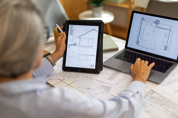 istock Older woman architect working with blueprints on tablet and laptop 2239490210