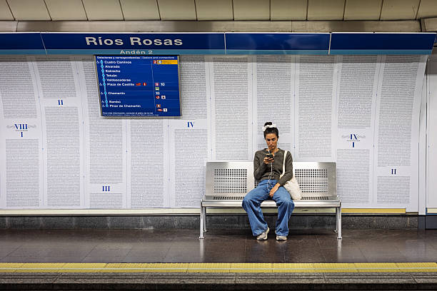 Rios Rosas station, Madrid, Spain, Europe, September 10, 2025:  A young woman sits alone on a metal bench at Rios Rosas metro station in Madrid, focused on her smartphone. She wears casual clothing and has a light blue bag beside her. The station’s wall behind her features a literary-themed design resembling printed book pages, adding cultural depth to the transit setting.