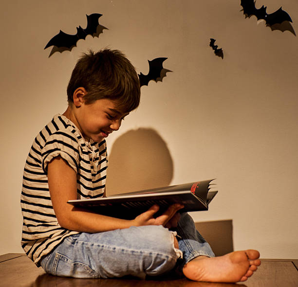 istock Enthusiastic child or smiling boy reading a book in cozy Halloween setting with bat decorations on wall. Warm lighting creates festive, spooky autumn atmosphere. Perfect for seasonal education themes 2239503322