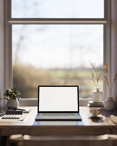 White screen laptop with calculator and coffee cup on wooden table under window in warm sunlight.