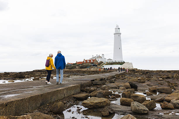 A wide shot of an elderly couple dressed in bright waterproof jackets walks hand-in-hand along a rocky coastal path toward a tall white lighthouse. The scene captures a peaceful seaside moment, with overcast skies and scattered rocks leading to the lighthouse surrounded by small houses. Perfect for themes of travel, retirement, companionship, and coastal exploration.Videos are available similar to this scenario.