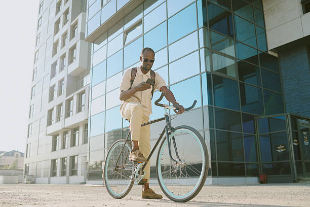 istock Young Adult Black Man Riding Bicycle While Using Smartphone near Office Building 2239507241