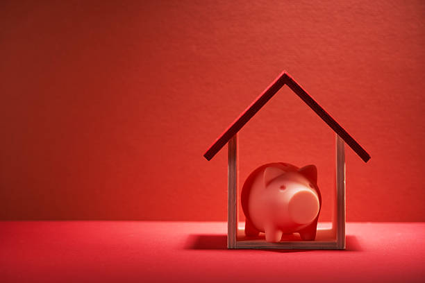 Stock photo showing a close-up view of a purple cardboard house with piggy bank money box on roof. This is a family finances, real estate and holiday savings concept picture.
