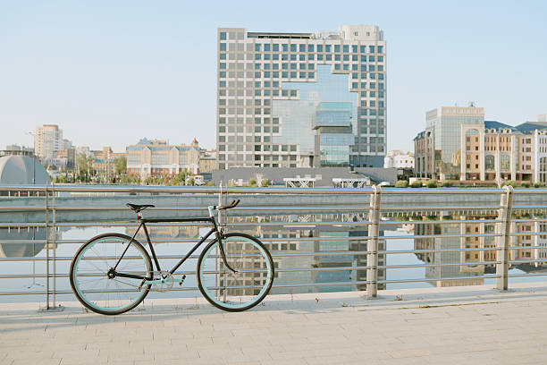 Single bicycle leaning against metal railing near waterfront with modern city buildings in background, reflecting in calm water, urban outdoor scene with copy space