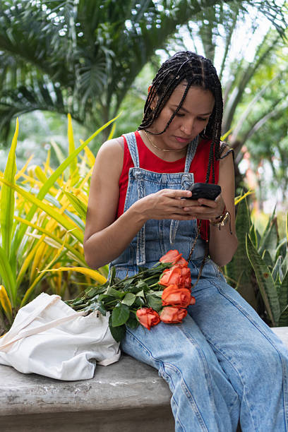 istock Woman with braids using smartphone relaxing in park with roses 2239516494