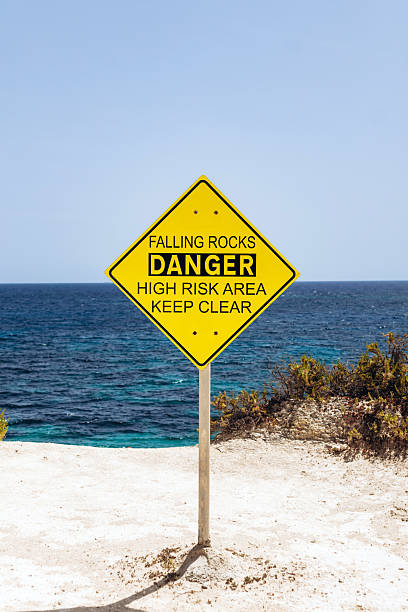 istock Bright yellow warning sign reading Falling Rocks  Danger High Risk Area Keep Clear stands on a sandy cliffside path with the deep blue Mediterranean Sea in the background 2239516802