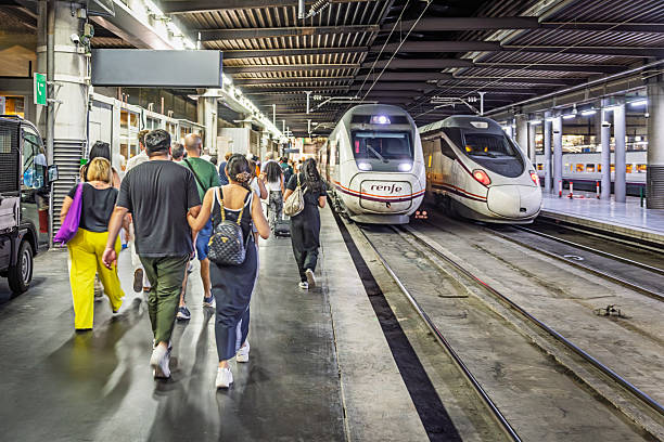 Atocha railroad station, Madrid, Spain, Europe, September 16, 2025:  Travelers walk along the platform at Atocha station in Madrid, next to a RENFE high-speed train with a sleek white exterior and red detailing. The station’s industrial ceiling, glass partitions, and overhead lighting frame a busy transit environment, as passengers carry luggage and prepare to board or disembark.