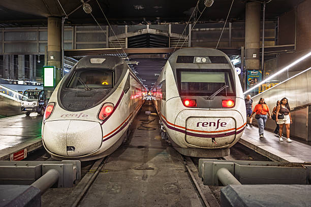 Atocha railroad station, Madrid, Spain, Europe, September 16, 2025:  Travelers walk along the platform at Atocha station in Madrid, next to a RENFE high-speed train with a sleek white exterior and red detailing. The station’s industrial ceiling, glass partitions, and overhead lighting frame a busy transit environment, as passengers carry luggage and prepare to board or disembark.