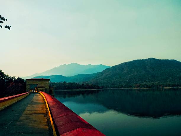 A long dam wall with a bright red boundary leads toward a small stone structure. Beyond lies a calm reservoir reflecting layered, misty hills under a pale sky.