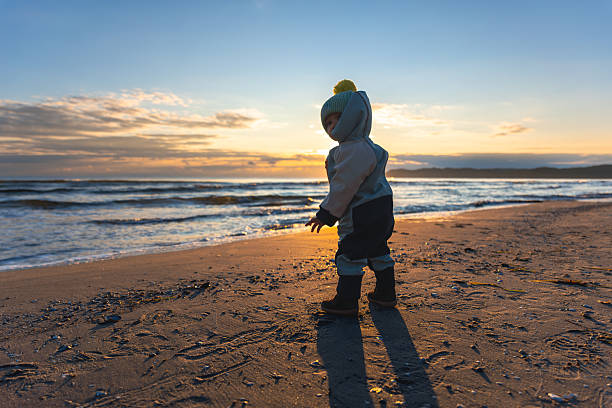 Child in warm clothes standing on sandy beach at sunrise. The long shadow and golden light create magical morning vibes of the coast.