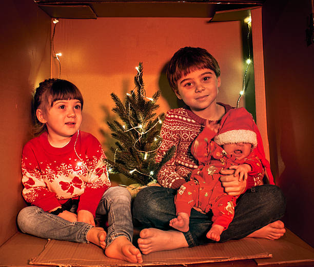Three children sit in a cozy cardboard box decorated with fairy lights and a tiny tree. Dressed in festive sweaters, they create a warm, DIY Christmas scene symbolizing joy, simplicity and family love