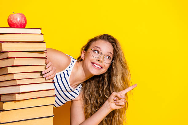 istock Young cheerful student next to a stack of book on a yellow background, pointing and showcasing education, learning 2239537792